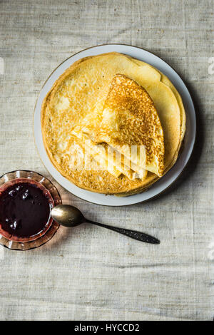 Pila di fogli piegati golden crepes sulla piastra bianca, nero Confettura di ribes in crystal rosetta, cucchiaio, tela di lino, vista dall'alto Foto Stock