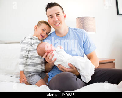 Padre che porta figlia (0-1 mesi) e seduto a letto con il figlio (4-5) Foto Stock