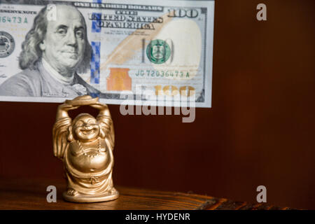 Un piccolo golden BUDDA STATUETTA detiene un centinaio di dollari in valuta degli Stati Uniti sopra la sua testa Foto Stock