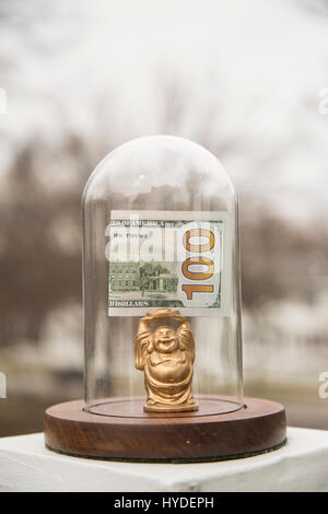 Un piccolo golden BUDDA STATUETTA in piedi in un display in vetro cupola contiene ripiegata di un centinaio di dollari sopra la sua testa Foto Stock