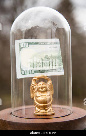 Un piccolo golden BUDDA STATUETTA in piedi in un display in vetro cupola contiene ripiegata di un centinaio di dollari sopra la sua testa Foto Stock