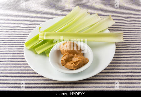 Vari gambi di sedano con una tazza di burro di arachidi su una piastra bianca sulla cima di un blu tovaglia a righe. Foto Stock