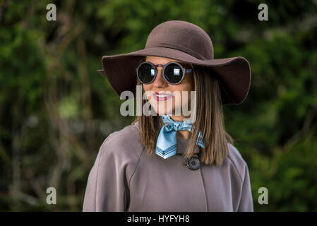 Giovane donna in hat, impermeabile e occhiali da sole è in posa nel parco durante una soleggiata giornata di primavera. Profondità di campo. Foto Stock