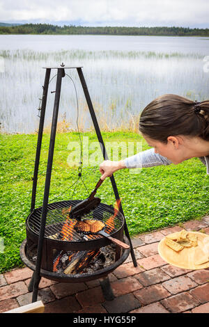 Giovane donna controllo di ghisa ferro waffle sul campo di fuoco, con pane appena sfornato waffle all'interno. Campeggio in natura, accanto al lago calmo. Foto Stock