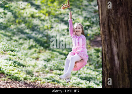 Carino bambina in abito rosa su albero swing corda in primavera in fiore giardino con fiori. Bambino oscillare all'aperto nel parco giochi del cortile. Bambini giocano ou Foto Stock