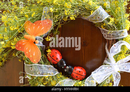 Ghirlanda di giallo fiori selvatici e decorate con una farfalla arancione e due di legno ladybugs su sfondo di legno Foto Stock