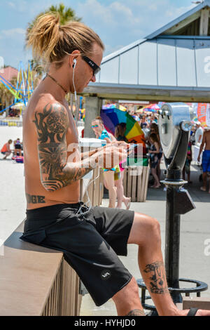 Beach bum con tatuaggi e le cuffie si siede sulle ringhiere di Pier 60 circondato da turista e fornitori con le persone che si godono i siti e la mercanzia lungo Foto Stock