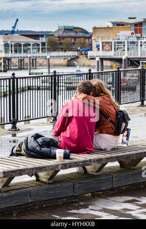 Un giovane seduto su una panchina e coccolare, affacciato sulla Baia di Cardiff, Cardiff Dock, nella primavera giornata piovosa, Cardiff Wales UK Foto Stock