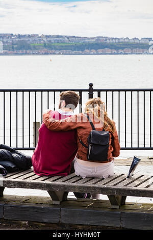 Un giovane seduto su una panchina e coccolare, affacciato sulla Baia di Cardiff, Cardiff Dock, nella primavera giornata piovosa, Cardiff Wales UK Foto Stock