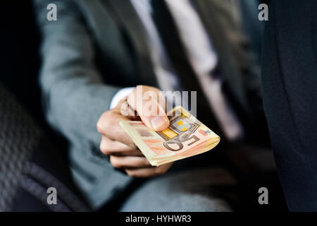 Un giovane uomo caucasico in un elegante abito grigio seduto nel sedile posteriore di una vettura offre un batuffolo di euro le fatture per la parte anteriore Foto Stock