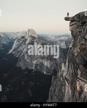 Un intrepido escursionista è in piedi su una roccia a strapiombo godendo della vista verso il famoso Half Dome al punto ghiacciaio nel Parco Nazionale di Yosemite in California Foto Stock