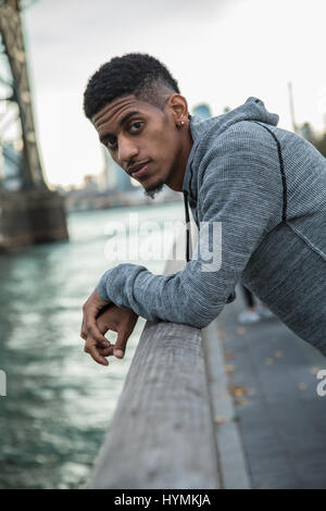 Un giovane uomo nero passeggiate lungo New York City East River con Brooklyn Williamsburg Bridge in background. Girato durante l'autunno del 2016. Foto Stock