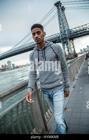 Un giovane uomo nero passeggiate lungo New York City East River con Brooklyn Williamsburg Bridge in background. Girato durante l'autunno del 2016. Foto Stock