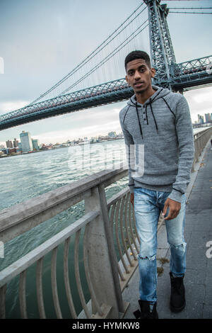 Un giovane uomo nero passeggiate lungo New York City East River con Brooklyn Williamsburg Bridge in background. Girato durante l'autunno del 2016. Foto Stock