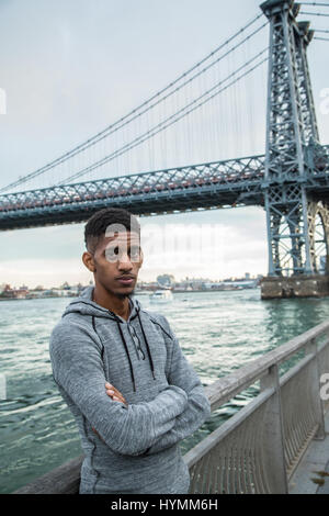 Un giovane uomo nero passeggiate lungo New York City East River con Brooklyn Williamsburg Bridge in background. Girato durante l'autunno del 2016. Foto Stock
