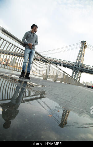 Un giovane uomo nero passeggiate lungo New York City East River con Brooklyn Williamsburg Bridge in background. Girato durante l'autunno del 2016. Foto Stock