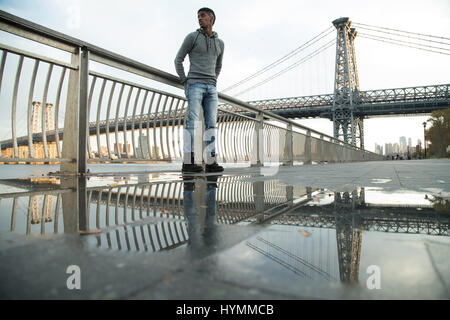 Un giovane uomo nero passeggiate lungo New York City East River con Brooklyn Williamsburg Bridge in background. Girato durante l'autunno del 2016. Foto Stock
