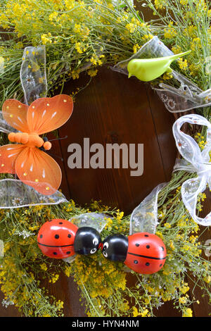 Ghirlanda di giallo fiori selvatici e decorata con ornamenti come una farfalla arancione verde piccolo uccello e due di legno ladybugs su sfondo di legno Foto Stock