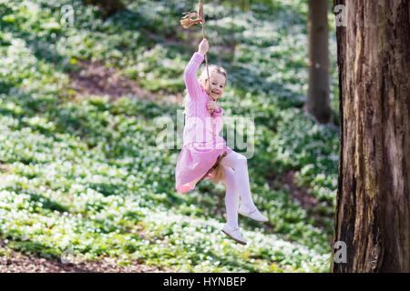 Carino bambina in abito rosa su albero swing corda in primavera in fiore giardino con fiori. Bambino oscillare all'aperto nel parco giochi del cortile. Bambini giocano ou Foto Stock