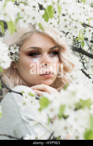 Bella ragazza con ricci capelli biondi nella primavera del giardino floreale, chiara buona giornata calda. Il tempo primaverile. Foto Stock