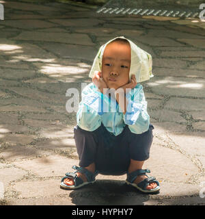 Giocoso cinese giovane ragazzo seduto su un percorso indossando giornale hat Foto Stock