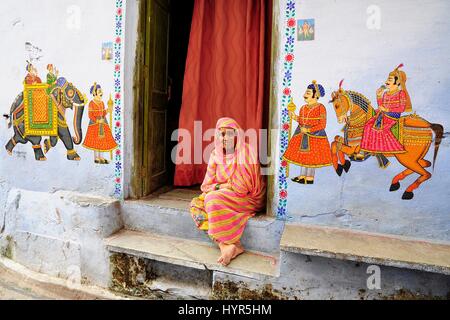 Udaipur, India, settembre 12, 2010: vecchia donna indiana seduto di fronte a lei huse su un terreno con dipinti su una parete. Foto Stock