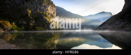 Straordinaria, fiaba, mitologici montagna lago paesaggio in una valle glaciale, con il sole che splende per il pendio roccioso e la nebbia di mattina al di sopra del lago Foto Stock