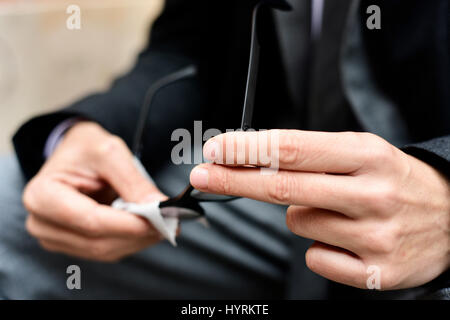 Primo piano di un giovane imprenditore caucasico in grigio di una tuta e un mantello nero pulizia delle lenti dei suoi occhiali con un panno in microfibra all'aperto Foto Stock