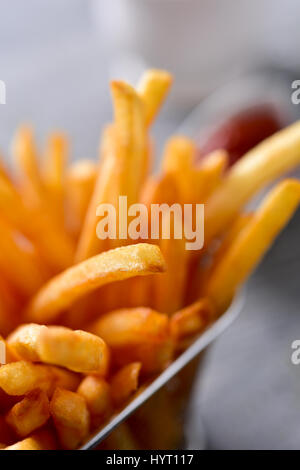 Primo piano di alcuni appetitosi patatine fritte servite in un cestello di metallo e alcune ciotole con la maionese e ketchup in background su un livello di grigio in legno rustico Foto Stock