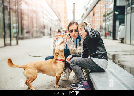 Madre mangiare panino e coccolare un cane mentre la figlia ascoltando musica su strada Foto Stock