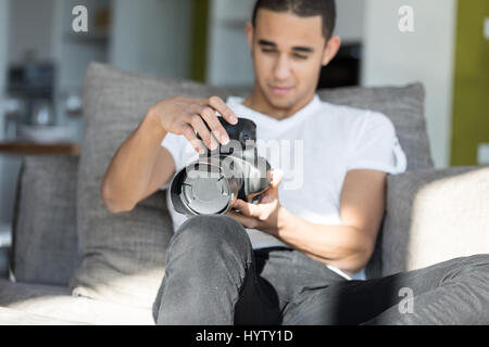 Vista frontale dei giovani brunette maschio bianco in maglietta e jeans seduti sul divano grigio interni tenendo la fotocamera DSLR e guardando lo schermo Foto Stock