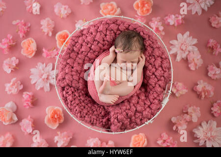 Nove giorni di età neonato ragazza dorme in un cesto in filo. Girato in studio su un sfondo rosa che è stato cosparso di fiori. Foto Stock