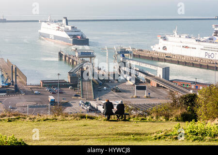 Inghilterra, Dover. Il primo piano è Edlerly giovane seduto su un banco di lavoro su una scogliera che si affaccia sulla car ferry terminal traghetti con inserito e la vela in background. Vista da dietro il giovane, distante shot. Foto Stock