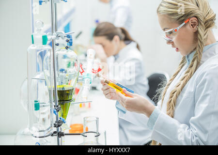 Giovani donne scienziato in occhiali di protezione e camice guardando le provette per la prova in laboratorio Foto Stock