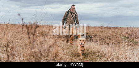 Escursionista e piccolo cane giallo a piedi nella prateria in un giorno nuvoloso Foto Stock