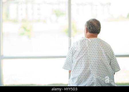 Paziente di sesso maschile che indossa camice guardando attraverso la finestra dell'ospedale, in vista posteriore Foto Stock