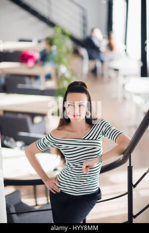 Ritratto di una donna bellissima guardando la telecamera vicino a una grande finestra. Adulto cafe proprietario con un sorriso accogliente negli interni moderni. Foto Stock