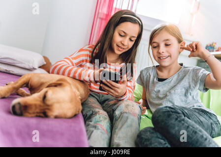 Due ragazze con small yellow dog sitter nella camera dei ragazzi e utilizzando smart phone Foto Stock