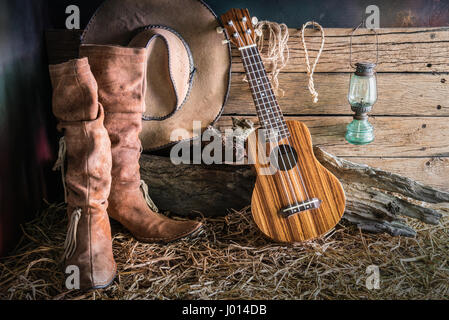 Still Life fotografia pittura con ukulele su american west rodeo feltro marrone cappello da cowboy e tradizionali stivali in pelle nel ranch vintage fienile backgrou Foto Stock