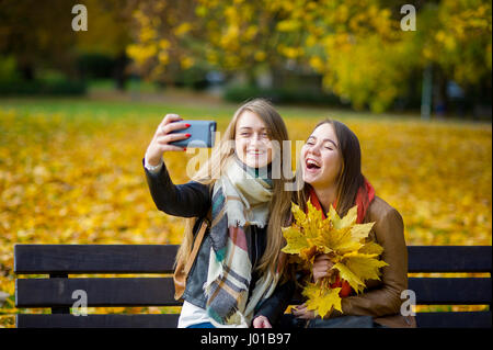 Mellow autunno. Due graziosi studenti fa un selfie nell'autunno del parco. Ragazze sedersi accanto gli uni con gli altri su una panchina nel parco. Una ragazza mantiene lo smartphone nel Foto Stock