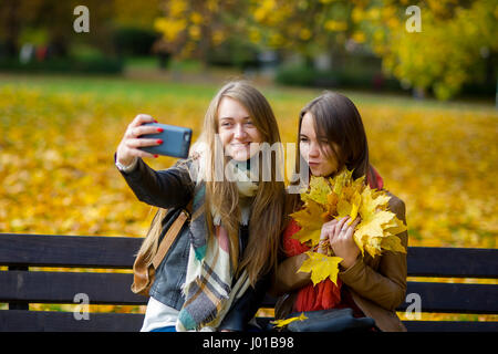 Mellow autunno. Due graziosi studenti fa un selfie nell'autunno del parco. Ragazze sedersi accanto gli uni con gli altri su una panchina nel parco. Una ragazza mantiene lo smartphone nel Foto Stock