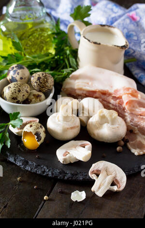 Sfondo - culinario su una scheda di ardesia di funghi, prosciutto, latte e uova di quaglia. Gli ingredienti freschi per la realizzazione di una frittata. Copia dello spazio. Foto Stock