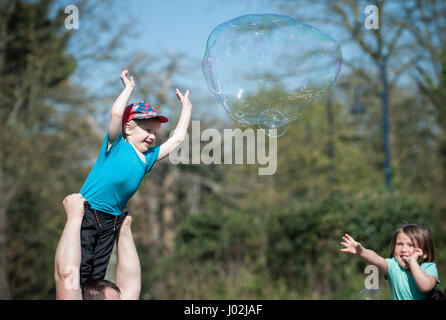 Un giovane ragazzo viene sollevato in aria da suo padre in modo che egli possa provare e burst grandi bolle galleggianti in aria durante una famiglia evento all'aperto nel parco di Cotam, Bognor Regis, West Sussex, Regno Unito. Foto Stock