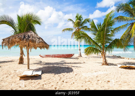 Gli alberi di palma, lettini per il vuoto, barca e l'ombrello sono sulla spiaggia di sabbia bianca. Mar dei Caraibi, Repubblica dominicana, Saona Island costa, località turistica Foto Stock