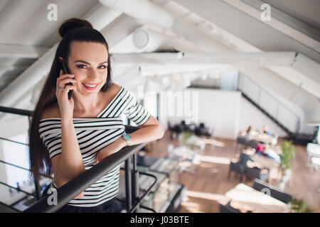 Ritratto di una donna in un cafe appoggiata contro la ringhiera e parlare con il suo telefono. Bella femmina adulta sorridente e guardando la telecamera. Foto Stock