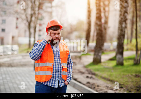 Ritratto di brutale barba lavoratore abito uomo lavoratore edile in arancio casco contro un marciapiede pensando. Foto Stock