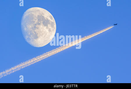 Le scie di condensazione (contrails) da un aereo jet con una luna quasi piena (a waxing gibbous), contro il cielo blu. Foto Stock
