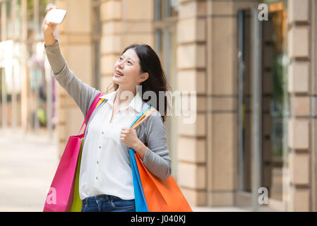 Shopping donna felice di prendere foto con lo smartphone. Donna che mantiene le borse della spesa tenendo Self-portrait immagine esterna. multirazziale donna asiatica Foto Stock