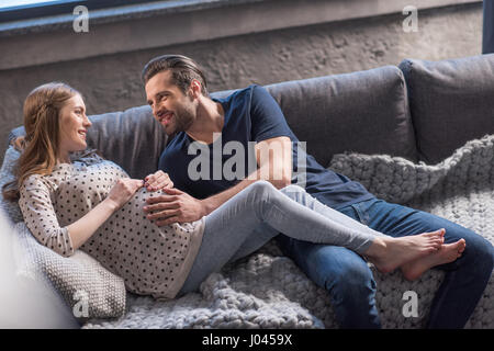 Uomo felice di toccare la pancia di sorridente donna incinta sdraiato sul divano Foto Stock