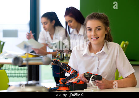 Ragazza adolescente in una progettazione e una tecnologia lezione. Lei è sorridente alla fotocamera con un braccio robotico che ella è la costruzione di fronte a lei. Foto Stock